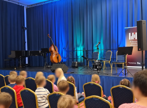 Na scenie z niebieskimi zasłonami występują artyści podczas koncertu. Widać fortepian, kontrabas, mikrofony, pulpity i krzesła dla muzyków oraz kolorowe oświetlenie sceniczne.