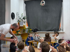 Dzieci siedzą przy stoliczkach i z zaciekawieniem obserwują świetlne doświadczenia. Na swoich twarzach mają specjalne okulary do obserwacji światła.