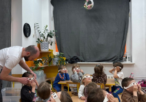 Dzieci siedzą przy stoliczkach i z zaciekawieniem obserwują świetlne doświadczenia. Na swoich twarzach mają specjalne okulary do obserwacji światła, dzięki którym mogą lepiej dostrzegać kolorowe efekty i niezwykłe zjawiska świetlne. Zajęcia wzbudzają w przedszkolakach dużo radości i zainteresowania.