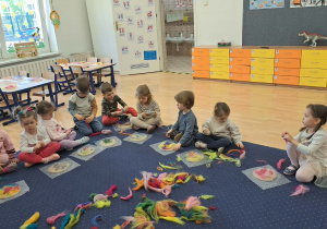 Dzieci z grupy Krasnale siedzą na dywanie podczas warsztatów filcowania. Przed nimi leży filc w różnych kolorach, a każde dziecko ma przed sobą swoją własną pracę, którą starannie przygotowuje. Przedszkolaki tworzą piękne prezenty dla mamy i taty w radosnej i twórczej atmosferze.