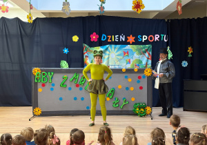 Dzieci oglądają przedstawienie na holu przedszkolnym. Na scenie występuje czarodziej i żaba.