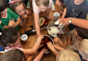 Przedszkolaki stoją wokół stolika z truskawkami, bananami i jogurtami. Dzieci wspólnie dodają składniki do garnka, przygotowując zdrowy koktajl owocowy.