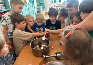 Przedszkolaki stoją wokół stolika z truskawkami, bananami i jogurtami. Dzieci wspólnie dodają składniki do garnka, przygotowując zdrowy koktajl owocowy.