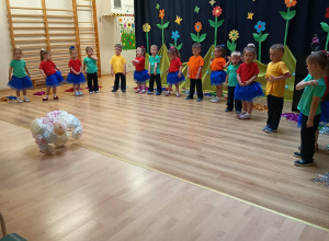 Przedszkolaki w kolorowych strojach stoją w półkolu podczas występu z okazji Dnia Mamy i Taty.