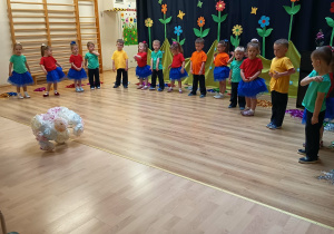 Przedszkolaki w kolorowych strojach stoją w półkolu podczas występu z okazji Dnia Mamy i Taty.