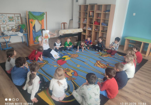 Dzieci siedzą w kółeczku na dywanie z panią prowadzącą.