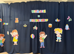 Granatowa kotara z napisem "Witajcie w przedszkolu" ozdobiona barwnymi postaciami dzieci i zabawkami.