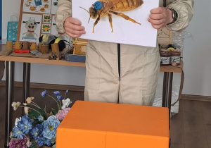 Mężczyzna w stroju pszczelarza stoi i pokazuje zdjęcie pszczoły. Ma na sobie ochronny kapelusz z siatką i opowiada o pracy pszczół.