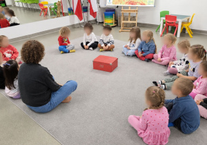 Dzieci siedzą na dywanie, w kręgu z panią prowadzącą zajęcia.