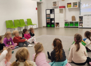 Przedszkolaki siedzą na dywanie i słuchają prowadzącej, a na tablicy interaktywnej wyświetlane są materiały edukacyjne.