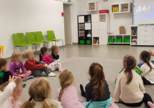 Przedszkolaki siedzą na dywanie i słuchają prowadzącej, a na tablicy interaktywnej wyświetlane są materiały edukacyjne.