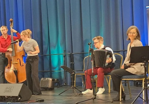 Na scenie z niebieskimi zasłonami występują artyści podczas koncertu. Widać fortepian, kontrabas, mikrofony, pulpity i krzesła dla muzyków oraz kolorowe oświetlenie sceniczne.