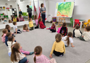 Grupa dzieci siedzi na dywanie i uczestniczy w warsztatach bibliotecznych. Na ekranie wyświetlana jest prezentacja przedstawiająca jego postać, a obok stoi mapa Polski.