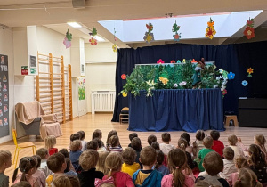 Na zdjęciu widać dzieci siedzące i oglądające teatr kukiełkowy. Przedstawienie odbywa się na scenie, a mali widzowie z zaciekawieniem śledzą akcję, uczestnicząc w wydarzeniu w radosnej i skupionej atmosferze.